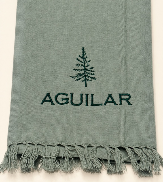 Sage-gray fringed towel embroidered with a dark green evergreen tree and AGUILAR.