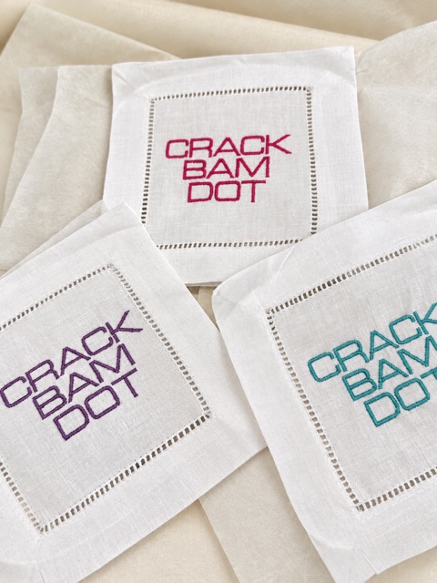 Three white hemstitch napkins with stacked CRACK BAM DOT embroidery in magenta, purple and teal.