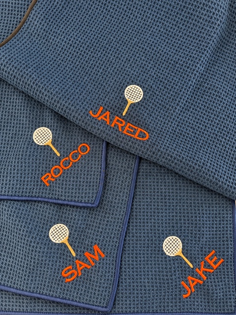 Four charcoal-blue tennis towels named JARED, ROCCO, SAM and JAKE in orange beneath cream racket motifs.