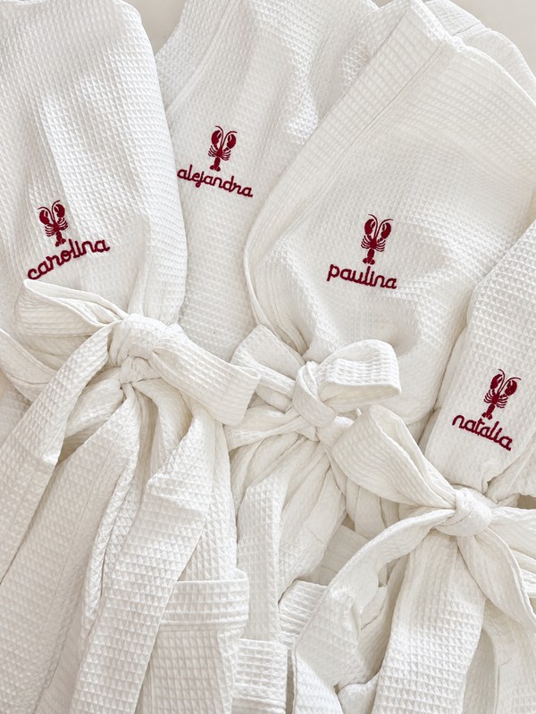 Four white waffle robes with burgundy lobster motifs and the names carolina, alejandra, paulina and natalia.
