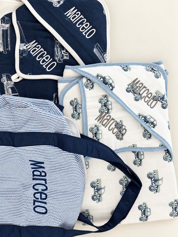 Navy and white vehicle-print baby towels and cloths embroidered Marcelo beside a matching personalized blue striped bag.