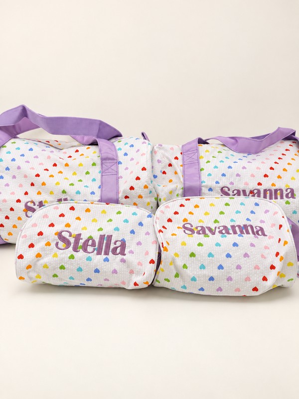 Two heart-print duffels behind matching zip cases embroidered Stella and Savanna in purple.