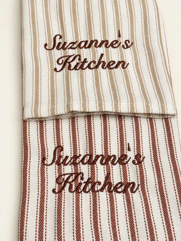 Two cream kitchen towels with beige and brown stripes and brown Suzanne’s Kitchen embroidery.